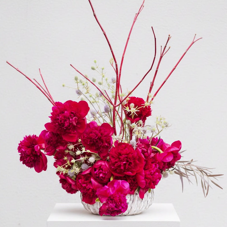 Floral arrangement with red Peony flowers and branches on a white pedestal against a light gray background