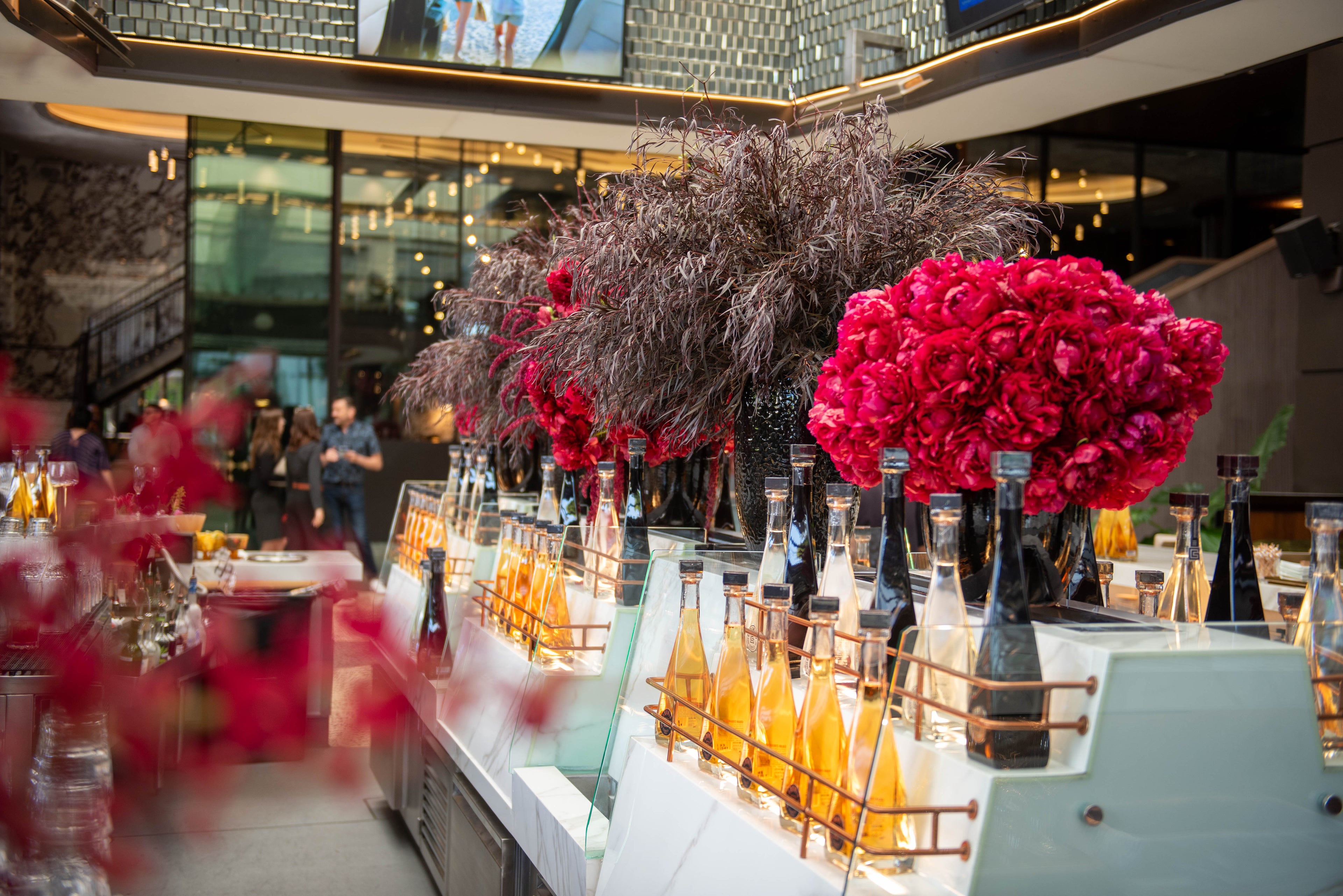 Lavish event beverage display with red peony arrangements and rows of champagne bottles — Beverly Hills Florist, Los Angeles