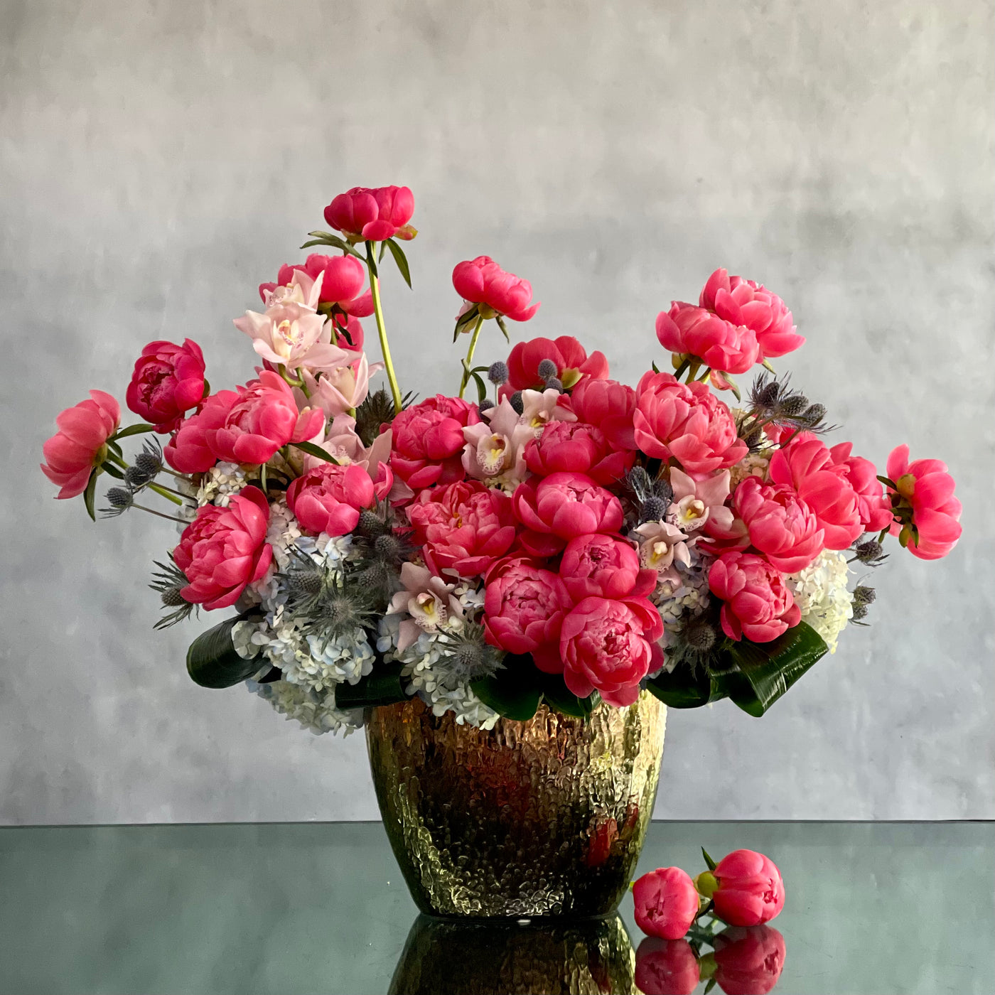 A luxurious floral arrangement featuring vibrant pink peonies and delicate white accents, displayed in a gold-toned vase with a reflective surface. The flowers are artistically arranged to create a lush, elegant centerpiece against a soft gray background