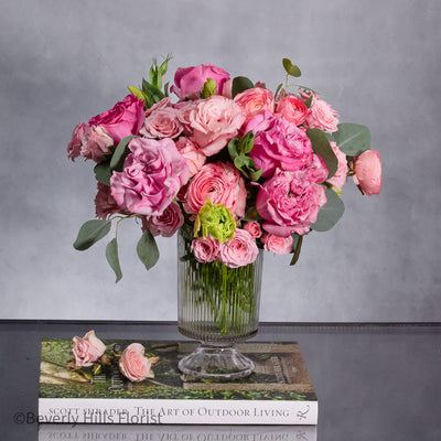 A Case of Hot Fever arrangement featuring fresh garden roses and ranunculus in a tall clear vase, designed in a circular formation, perfect for all occasions.