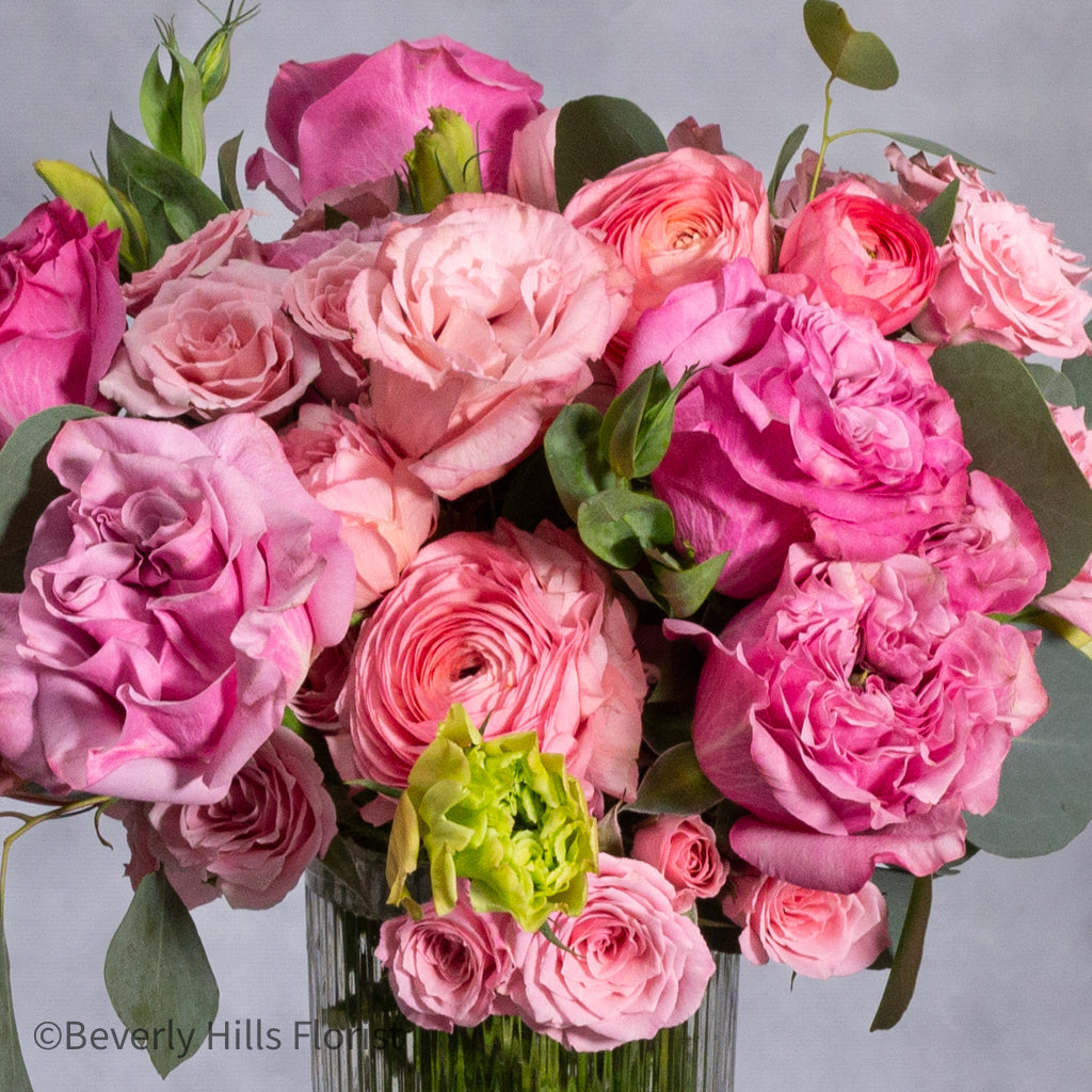 A Case of Hot Fever arrangement featuring fresh garden roses and ranunculus in a tall clear vase, designed in a circular formation, perfect for all occasions.