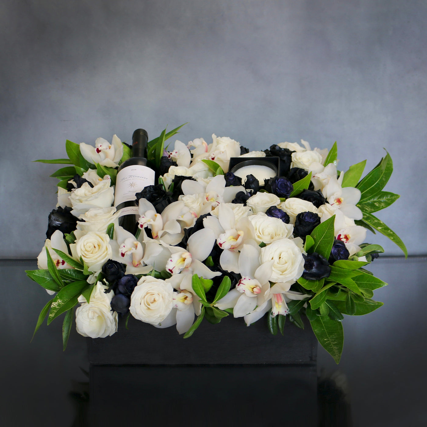 A Gift For You arrangement with black and white roses, cymbidium orchids, greens in a black wooden box with wine and a candle, by Beverly Hills Florist.