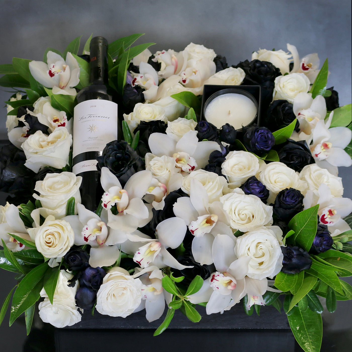 A Gift For You arrangement with black and white roses, cymbidium orchids, greens in a black wooden box with wine and a candle, by Beverly Hills Florist.