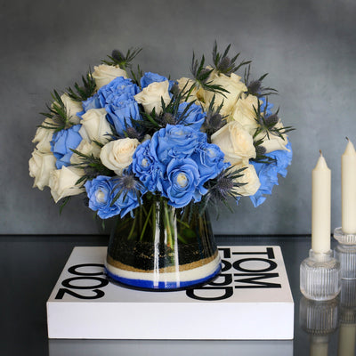 Blue Me Away arrangement with blue and white roses, thistles, and colored sand in a modern vase, available for same-day delivery by Beverly Hills Florist.