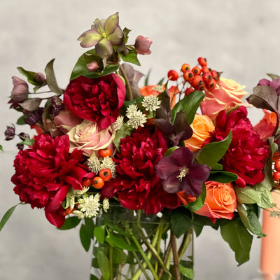 Majestic winter arrangement with orange roses, burgundy peonies, and winter accents in a glass vase – available for same-day delivery by Beverly Hills Florist.