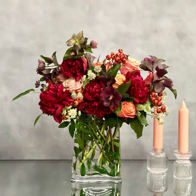 Majestic winter arrangement with orange roses, burgundy peonies, and winter accents in a glass vase – available for same-day delivery by Beverly Hills Florist.