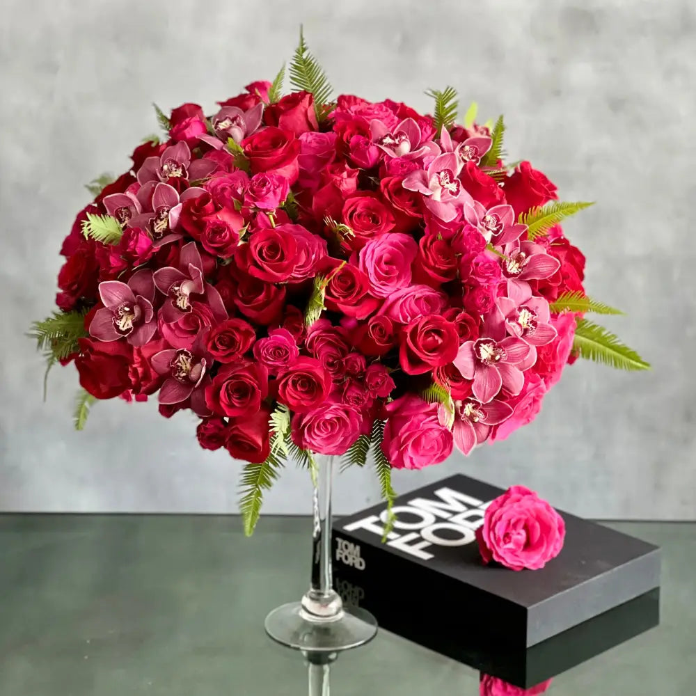 Elegant floral arrangement with red and pink roses, burgundy cymbidium orchids, and greens in a tall pedestal glass vase