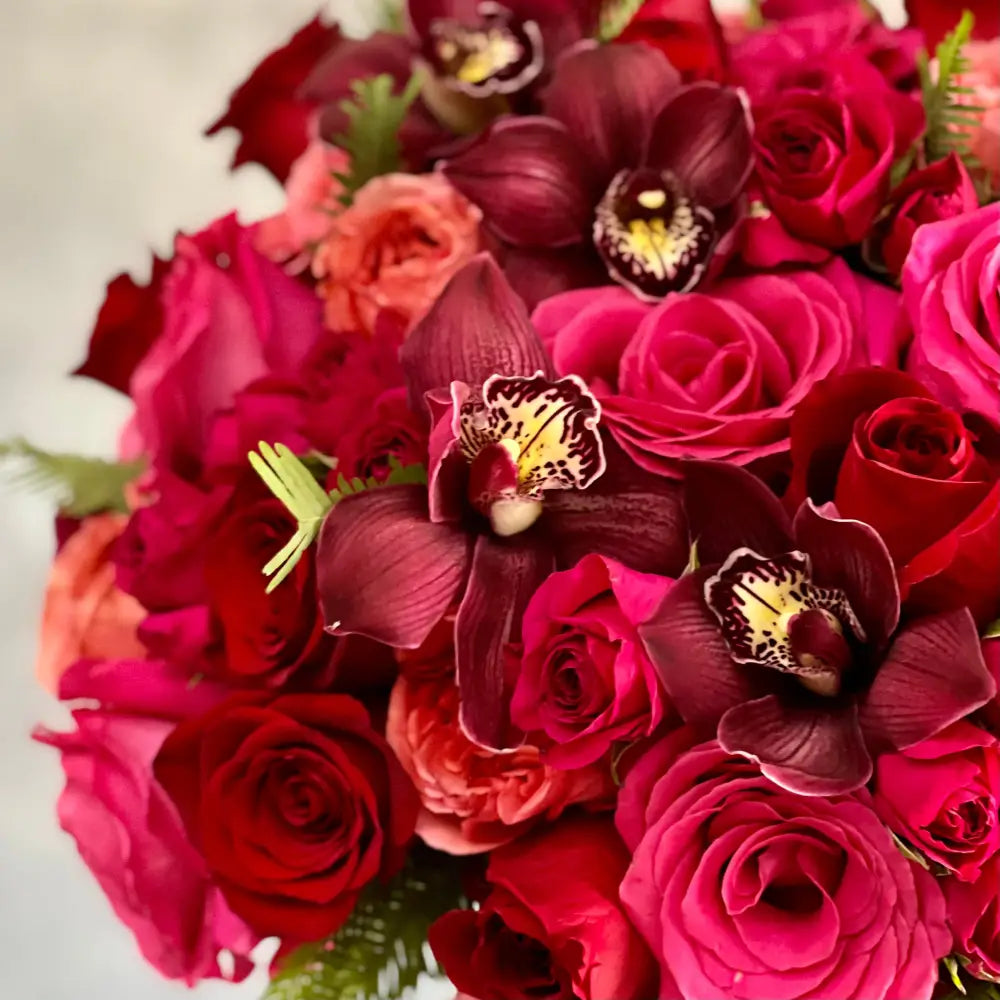 Elegant floral arrangement with red and pink roses, burgundy cymbidium orchids, and greens in a tall pedestal glass vase