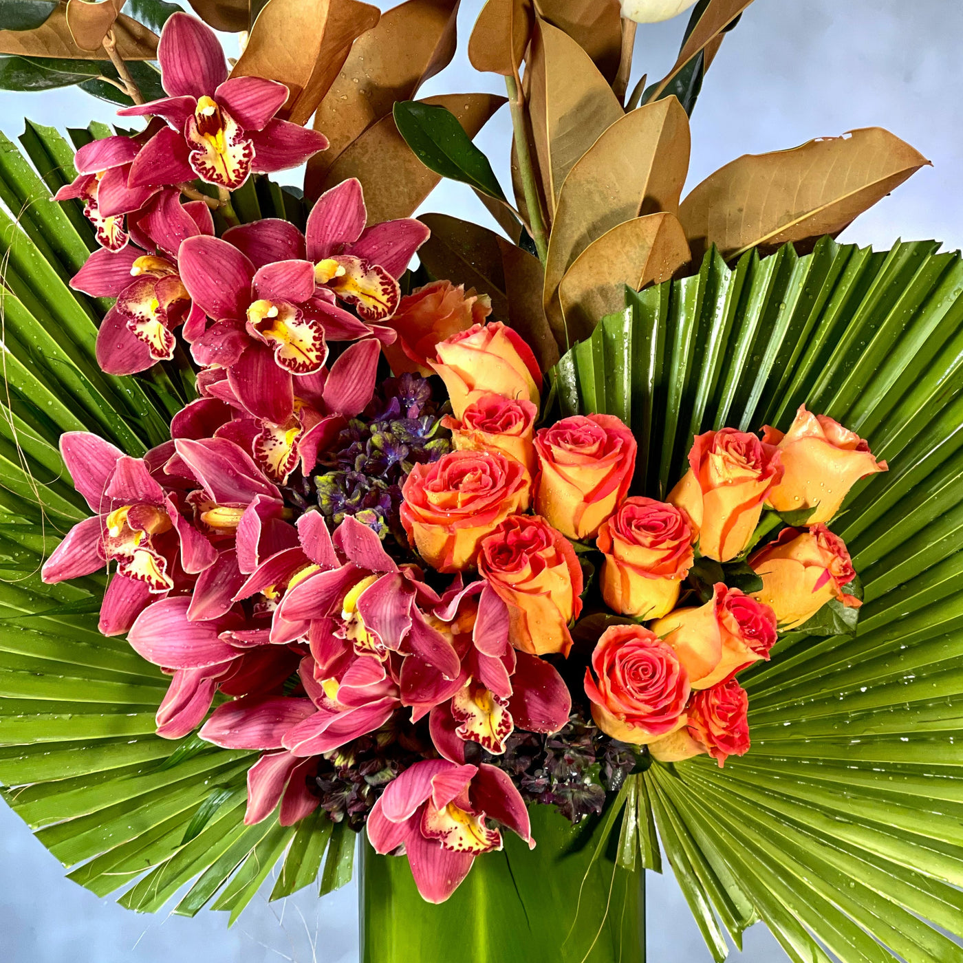 A Dream Come True arrangement with orange roses, cymbidium orchids, and fan palms in a tall glass vase, same-day delivery by Beverly Hills Florist.