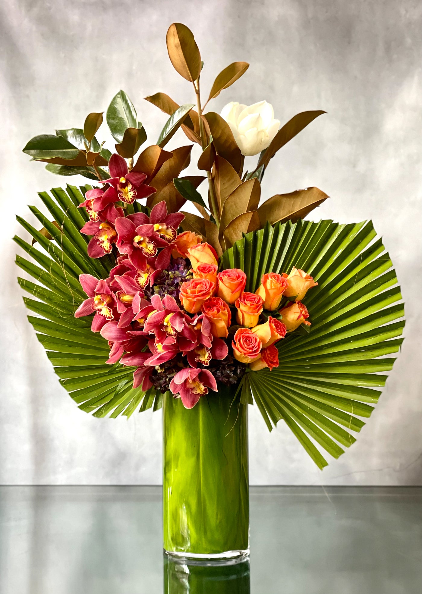 A Dream Come True arrangement with orange roses, cymbidium orchids, and fan palms in a tall glass vase, same-day delivery by Beverly Hills Florist.