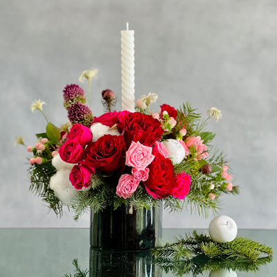 Festive Christmas centerpiece with red and pink roses, seasonal greens, berries, and glowing tapers, approx. 14 inches tall, for same-day delivery.