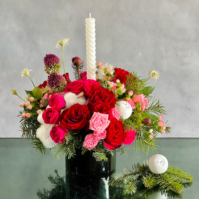 Festive Christmas centerpiece with red and pink roses, seasonal greens, berries, and glowing tapers, approx. 14 inches tall, for same-day delivery.