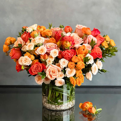 Elegant floral arrangement with orange roses, peach spray roses, ranunculus, and greenery in a round 6x6 clear glass vase.