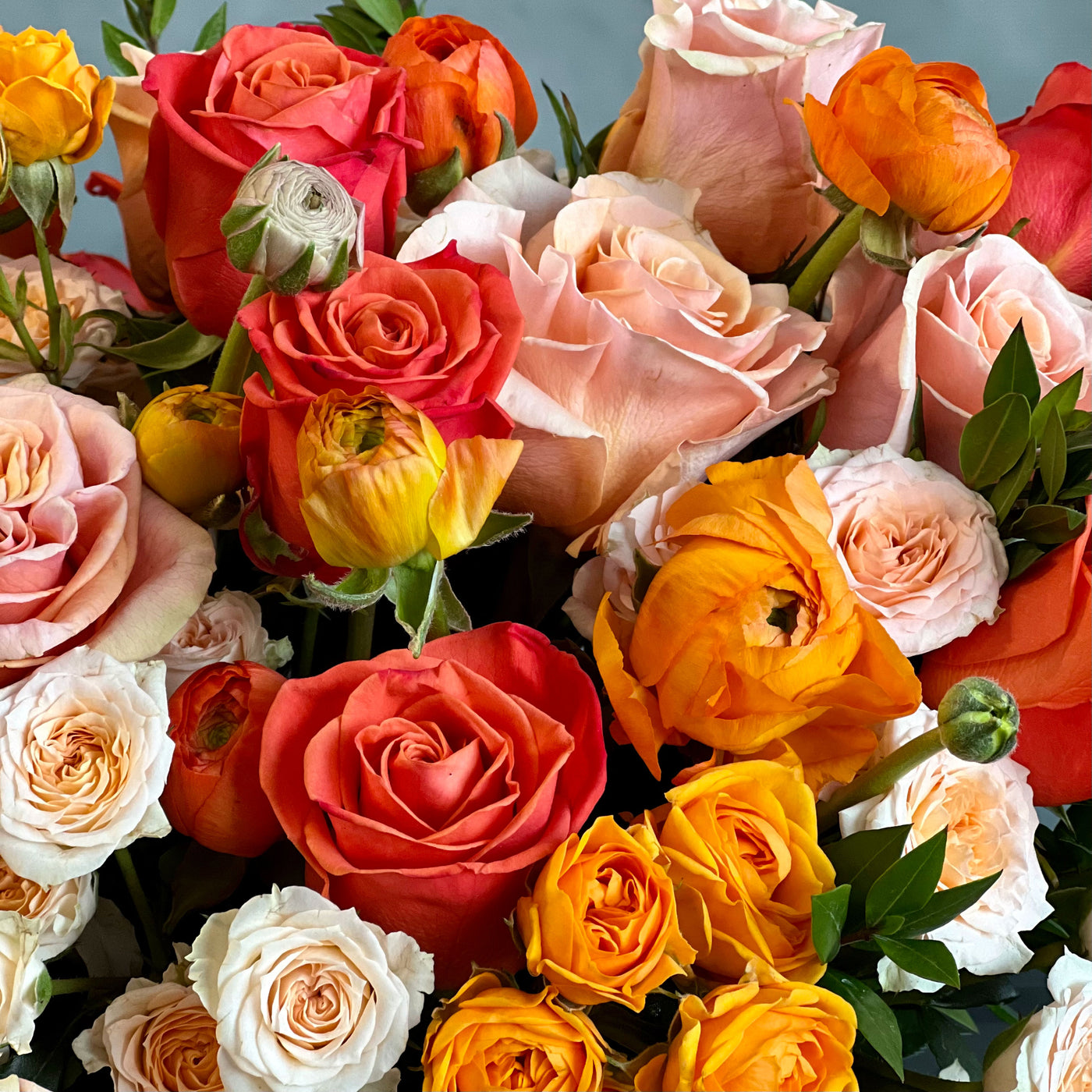 Elegant floral arrangement with orange roses, peach spray roses, ranunculus, and greenery in a round 6x6 clear glass vase.