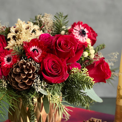 Yuletide Spirit holiday floral arrangement with red roses, spray roses, natural accents, and a gold ornament in a brushed gold cylinder vase.